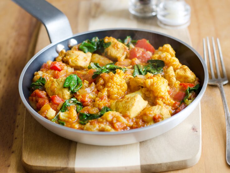 Schmackhaftes Hühnchen-Curry mit Tomaten und Spinat, serviert in einer Pfanne auf einem Holzbrett.