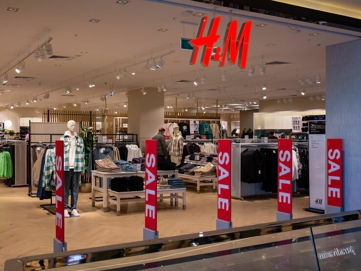 Ein H&M-Bekleidungsgeschäft mit gut beleuchtetem, geräumigem Innenraum. Verschiedene Kleidungsstücke sind auf Mannequins und in Regalen ausgestellt. Auffällige rote "SALE"-Schilder sind im Vordergrund zu sehen, die auf Sonderangebote hinweisen. Im Hintergrund ist das charakteristische H&M-Logo in roten Buchstaben deutlich sichtbar. Der Laden scheint ruhig, ohne viele Kunden, was eine entspannte Einkaufsatmosphäre schafft.